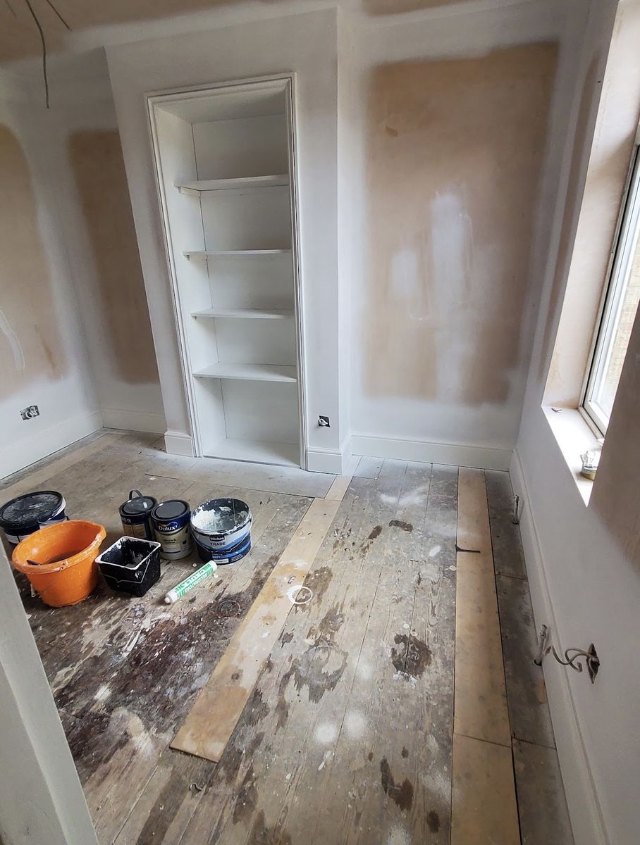 A room in bare pink plaster with alcove shelving primed white and paint tins on a bare floor