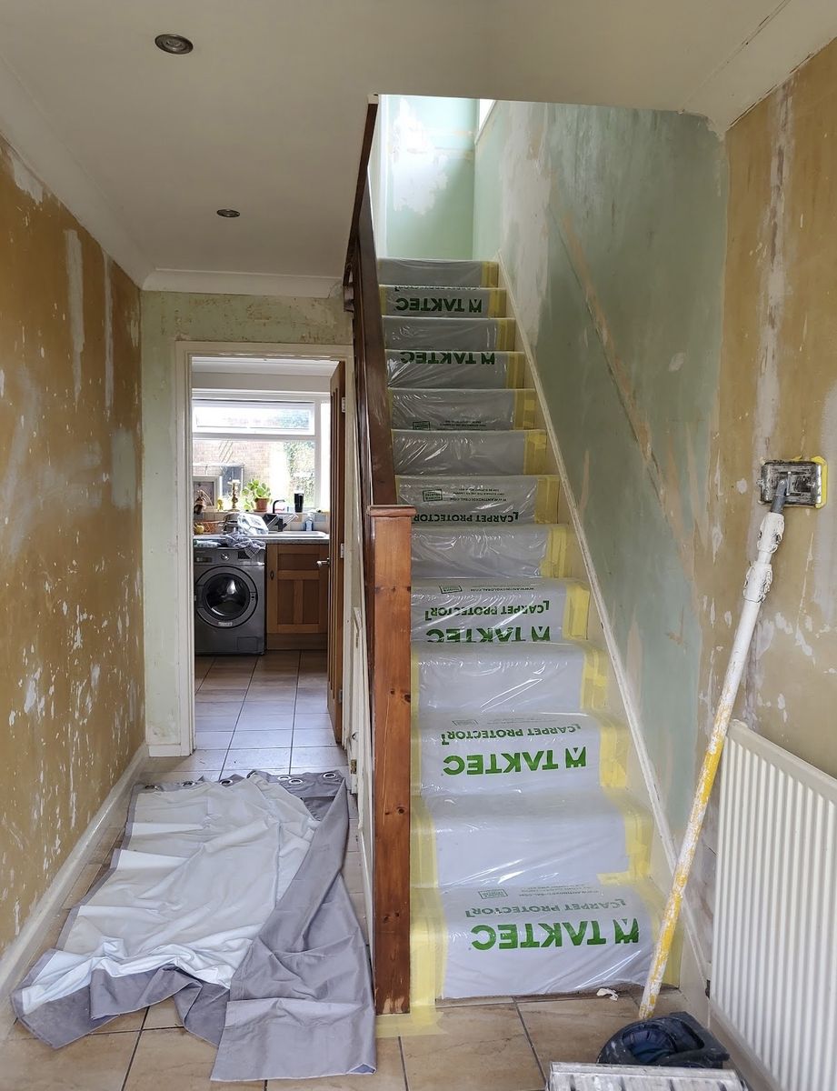 A hallway stripped back to patchy bare plaster, stair treads covered with protective sheeting